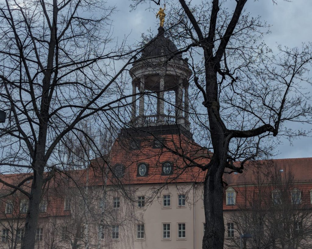 Ein Bild des Großen Waisenhauses in Potsdam, verdeckt durch kahle Äste eines vordergründigen Baumes. Der Himmel ist bewölkt, und das historische Gebäude zeigt einen markanten Turm mit einer Kuppel und einer glänzenden Wetterfahne. Die Architektur des Waisenhauses ist im barocken Stil gehalten, mit einem roten Ziegeldach und hell verputzten Wänden.
